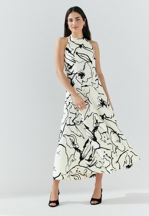 Woman with long dark hair wears sleeveless, white halter dress with abstract black floral print, paired with black open-toe heels, standing against plain background.