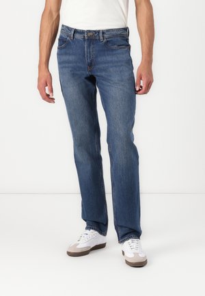 Jean en denim bleu pour homme avec une coupe droite, présentant un design classique à cinq poches et des détails de couture subtils. Porté avec des baskets blanches.