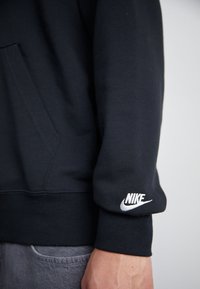Sweat à capuche noir avec une poche frontale, des poignets côtelés et un accent du logo Nike blanc sur la manche. Tissu lisse avec une coupe décontractée.