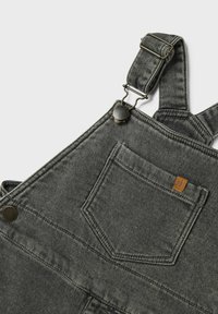 Salopette en denim gris avec des boucles en métal ajustables, poches latérales et une poche arrière. Tissu texturé avec des détails de couture minimalistes.