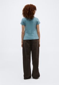 Γυναίκα με κατσαρά μαλλιά που φοράει ανοιχτό μπλε κοντομάνικο πουκάμισο και άνετο σκούρο καφέ παντελόνι, στέκεται πλάτη σε λευκό φόντο.