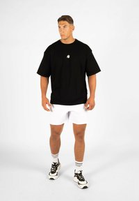 T-shirt noire oversize avec un petit logo blanc, associée à un short blanc et des sneakers noires. Le modèle se tient devant un fond uni.