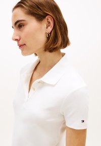 Polo blanc avec un col classique, doté de boutons dorés et de manches courtes. Petit logo discret sur la manche. Tissu doux et texturé.