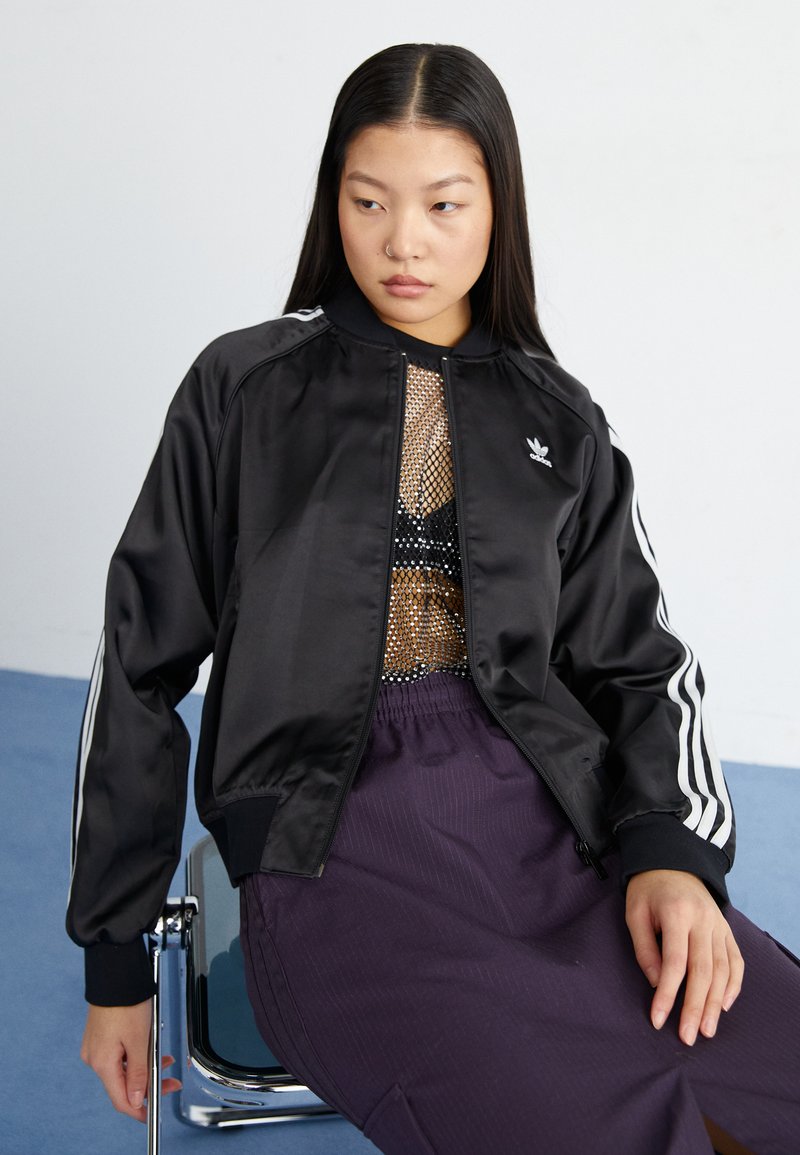 Black satin bomber jacket with white stripes, zippered front, ribbed cuffs, and collar. Worn over a mesh top and purple pants, seated on a chair.