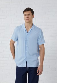 Chemise à manches courtes bleu clair en tissu léger, avec un devant boutonné et un col ouvert, accompagnée d'un pantalon sombre.