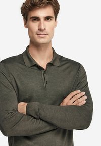 Mörkgrön långärmad polo med två knappar, ribbad krage och slät yta. Modellen står med armarna i kors.