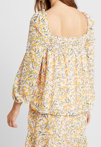 Kukkakuvioinen pusero puff-hihoilla, jossa on kootut takapaneelit ja vaaleanpunainen tausta, joka on koristeltu keltaisilla ja vihreillä lehtikuviolla.