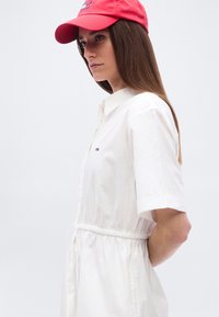 Γυναίκα με λευκό φόρεμα με κουμπιά και ρυθμιζόμενο κορδόνι στη μέση, φορά κόκκινο καπέλο με λογότυπο, στέκεται μπροστά σε απλό φωτεινό φόντο, κοιτώντας προς τα αριστερά.