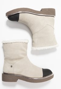 Botas de tobillo de ante beige con puntera ovalada negra, borde de piel blanca y suela marrón texturizada, con un detalle plateado a lo largo de la base.