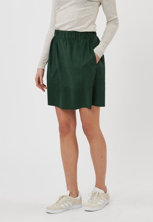 Jupe verte, mi-longue, avec une taille élastique, dotée de deux poches latérales et d'une texture lisse. Portée avec un haut à manches longues léger et des baskets.