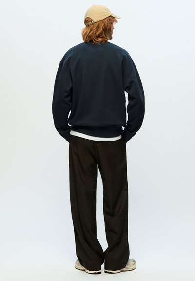 Sweatshirt marine avec des épaules tombantes et des poignets côtelés, porté par-dessus une couche blanche ; associé à un pantalon ample marron foncé et une casquette beige.