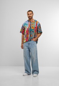 Mann steht in weit geschnittenen hellblauen Jeans, weißen Sneakers und einem lockeren, bunt gemusterten Kurzarmhemd mit einer Hand in der Tasche.