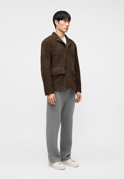 Άνδρας που φοράει καφέ σουέντ μπουφάν, γκρι παντελ όνι με φαρδιά μπατζάκια και λευκά αθλητικά, στέκεται μπροστά σε απλό φωτεινό φόντο.