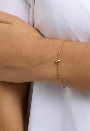 Hieno kultainen ketjurannekoru, jossa pieni sydänriipus, ranteessa valkoisen lyhythihaisen paidan päällä.