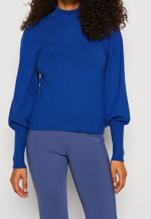 Femme portant un pull bleu vif à manches longues avec des manches bouffantes et des poignets ajustés, associé à un pantalon bleu assorti.