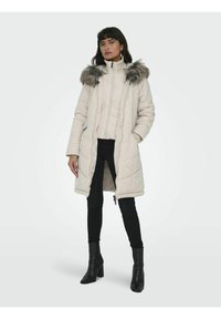 Manteau beige rembourré avec un col en fausse fourrure, doté d'un zip à l'avant, de poches latérales et d'une longueur jusqu'à mi-thigh, accompagné de jeans skinny noirs et de talons.