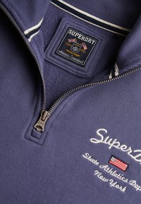 Pull Superdry bleu marine avec fermeture quart de zip, logo brodé, drapeau américain et texte "State Athletics Dept New York" sur la poitrine.
