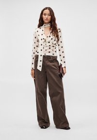 Biała bluzka z długim rękawem w brązowe groszki z wiązaniem, połączona z brązowymi szerokimi spodniami z wysokim stanem oraz małą ciemną torebką.