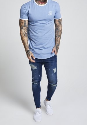 Mężczyzna z tatuażami na całym ramieniu, ubrany w jasnoniebieski dopasowany t-shirt, podarte obcisłe jeansy i białe trampki, stojący na jednolitym tle.