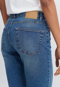 Detailní záběr na osobu v modrých džínách a bílém tričku, ukazující zadní kapsu a hnědou koženou nášivku.