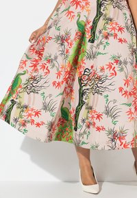 Robe midi florale en tissu doux avec un fond rose, ornée de paons verts, de feuilles rouges et de branches noires complexes. Portée avec des talons blancs.