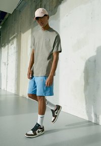 Mladý muž v růžové čepici, šedém tričku, modrých šortkách, bílých ponožkách Nike a hnědo-černých teniskách Nike, stojící proti texturované betonové zdi.