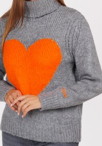 Vrouw in een grijze gebreide coltrui met een groot oranje hart op de borst en oranje borduursel op de mouw.
