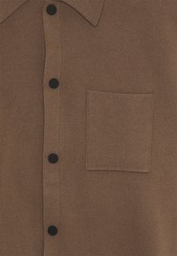 Chemise marron en tricot avec un col, dotée de boutons noirs et d'une poche carrée sur la poitrine. Texture lisse, design simple sans motifs.