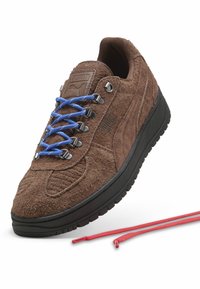 Brūni zamšaini sporta apavi ar ziliem šņorēm, sudraba acu caurumiem un melnu gumijas zoli. Blakus apaviem ir sarkanas aizvietojošas šņores.