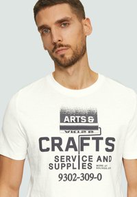 Witte katoenen T-shirt met een grafische print in donkergrijze tekst die leest "KUNST & HANDWERKDIENSTEN EN BENODIGDHEDEN" met een ontwerp van een verfroller.