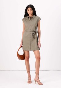 Abito camicia verde oliva con maniche corte, bottoni frontali e cintura legata in vita. Abbinato a sandali beige con cinturini e una borsa marrone.