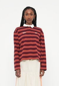 Polo majica z dolgimi rokavi z maroon in rjavimi oranžnimi črtami, belim ovratnikom in vezenim logotipom na prsih. Bombažni material, sproščen kroj.