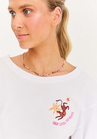 Blondýnka v bílém tričku s hnědým kočkou, hvězdicí a textem "Devět životů, žádná pravidla" a barevným korálkovým náhrdelníkem.