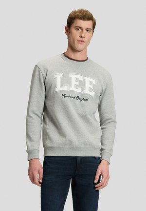 Uomo che indossa una felpa grigio chiaro con testo "LEE American Original", jeans scuri, rivolto in avanti contro uno sfondo grigio chiaro semplice.