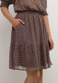 Žena v hnědých šatech s růžovým paisley vzorem, elastickým pasem a bočními kapsami, stojící proti světle šedému pozadí.