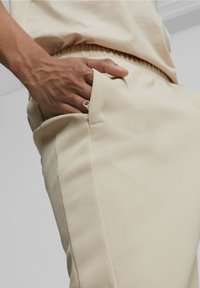 Beige texturerade byxor har ett elastiskt midjeband och sidofickor. En hand vilar i vänstra fickan, vilket framhäver plaggets passform.