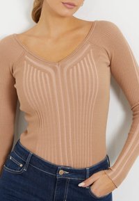 Langärmliges, geripptes Oberteil in hellem Beige mit einem V-Ausschnitt. Strukturierten, vertikalen Streifen und taillierter Silhouette. Kombiniert mit dunkelblauen Jeans.