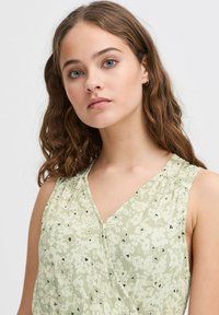Top sans manches à motifs floraux de couleur vert clair, avec un col en V, une texture subtile et de petites touches noires. Tissu doux, coupe décontractée.