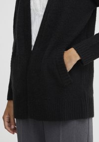 Svart stickad cardigan med öppen front, ribbade ärmslut och nederkant. Har sidofickor och ett mjukt, texturerat tyg. Bärs över grå byxor.