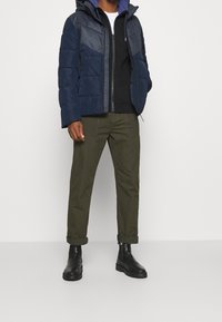 Doudoune bleu marine avec un panneau gris, fermeture éclair devant et capuche. Portée sur un pull noir et un pantalon vert olive, accompagnée de bottes noires.