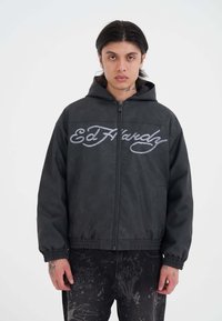 Giovane con lunghi capelli scuri che indossa una felpa nera con zip con il testo "Ed Hardy" e jeans scuri a fantasia, rivolto in avanti su uno sfondo bianco.