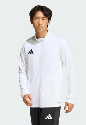 Młody mężczyzna w białej kurtce Adidas na zamek i czarnych spodniach Adidas, stojący z luźno opuszczonymi ramionami na jednolitym tle.