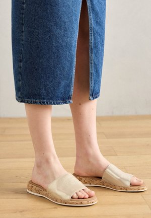Voeten met beige instapsandalen met kurken zolen, gecombineerd met een blauwe denim rok met een achterrits, op een houten vloer.