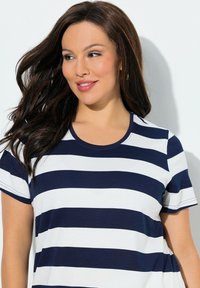 Frau mit langen dunklen Haaren, die ein kurzärmeliges, marineblau-weiß gestreiftes Shirt trägt, schaut leicht zur Seite und lächelt.