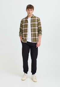 Menns rutete skjorte i olivengrønn og beige, knappe igjen, med en hvit T-skjorte med rund hals under, kombinert med svarte cargo-bukser og hvite joggesko.