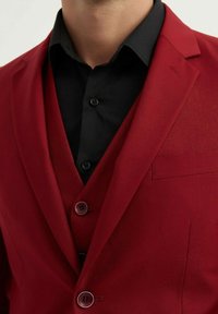 Blazer rouge au tissu lisse, fermeture à un bouton, et col cranté. Porté par-dessus une chemise noire à boutons.