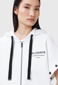 Sudadera blanca con cremallera y cordones negros, logo impreso, mangas cortas y acentos negros en los puños. Tejido suave, diseño casual.
