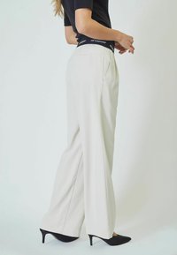 Wide-leg trousers in light beige fabric with a smooth texture. Features side pockets and a black branded waistband. Black pointed heels.