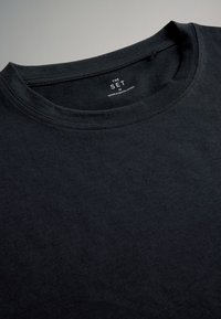 Schwarzes T-Shirt mit Rundhalsausschnitt und sichtbarem Innenetikett mit der Aufschrift „THE SET M Made in Bangladesh“ auf neutralem grauem Hintergrund.