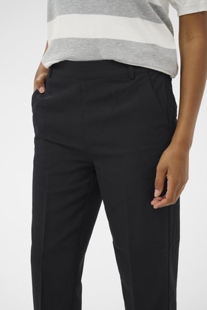 Asmuo dėvintis juodas pritaikytas kelnes ir pilkai baltai dryžuotą trumpomis rankovėmis marškinius, viena ranka kišenėje, kita atpalaiduota.